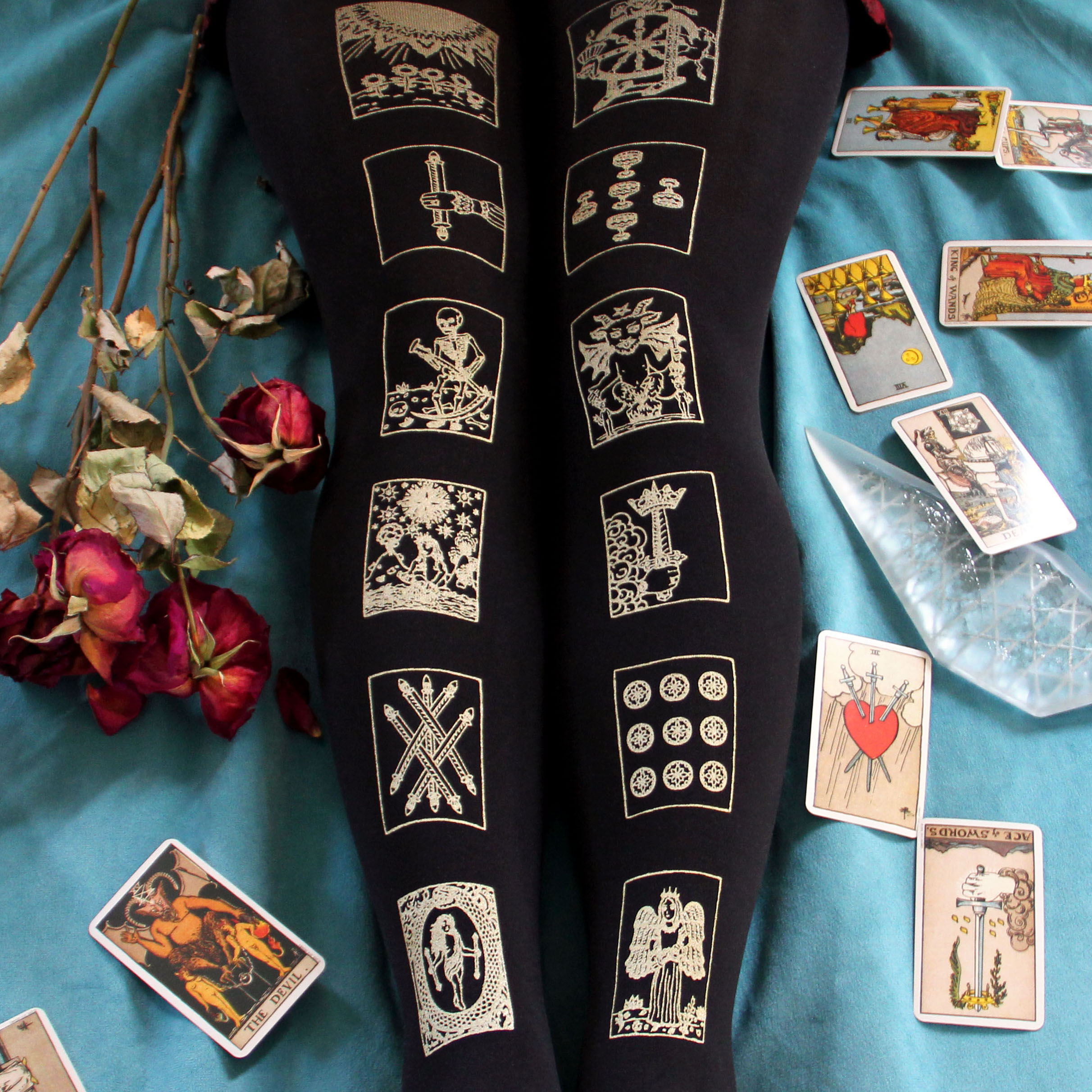 Screen printed tights with a design of tarot cards printed in gold, the design runs down the centre of each leg on black tights. There is a turquoise velvet background with red roses and tarot cards scattered around the legs.