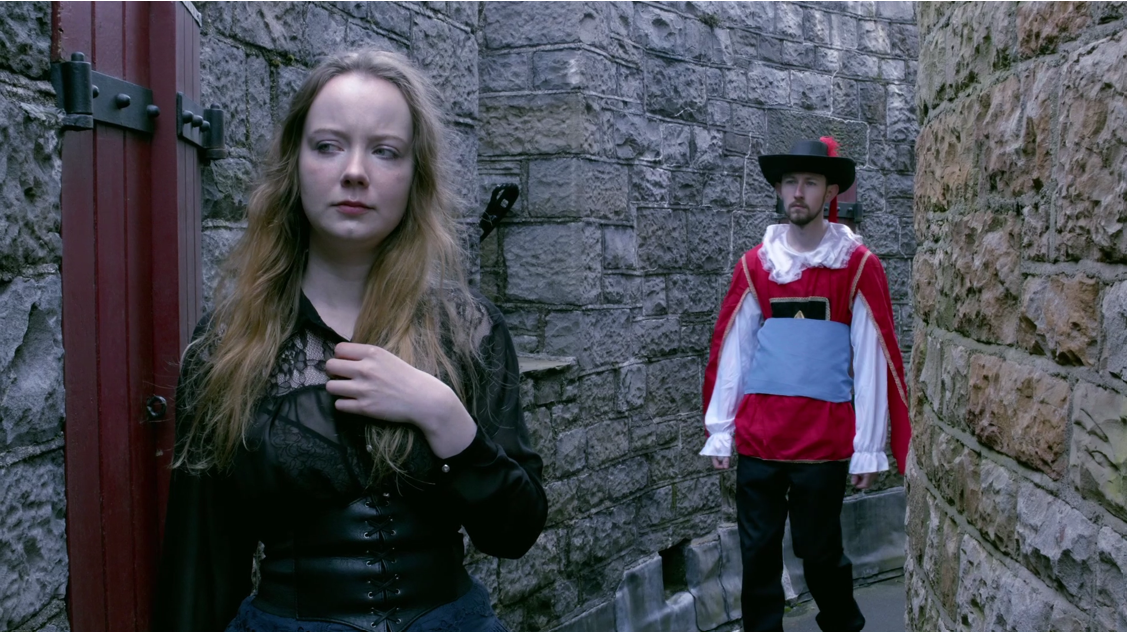 A young woman dressed in all-black Victorian / Edwardian upper class clothing stops in her tracks whilst walking along the turret of a castle, sensing someone behind her. To her right, on the bend of the turret, stands a man dressed in an English Civil War cavalier's uniform. He stares directly at her, his face emotionless.
