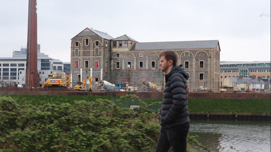 A young man walks along a riverbank. Behind him in the frame, we see an old industrial landscape on the other side of the river, complete with a large old brick factory building and chimney. A bulldozer can be seen on the site, and there are signs of redevelopment taking place.