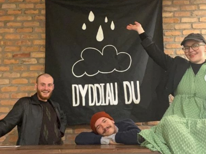The image features 3 members of Dyddiau Du around a table smiling with the logo in the back