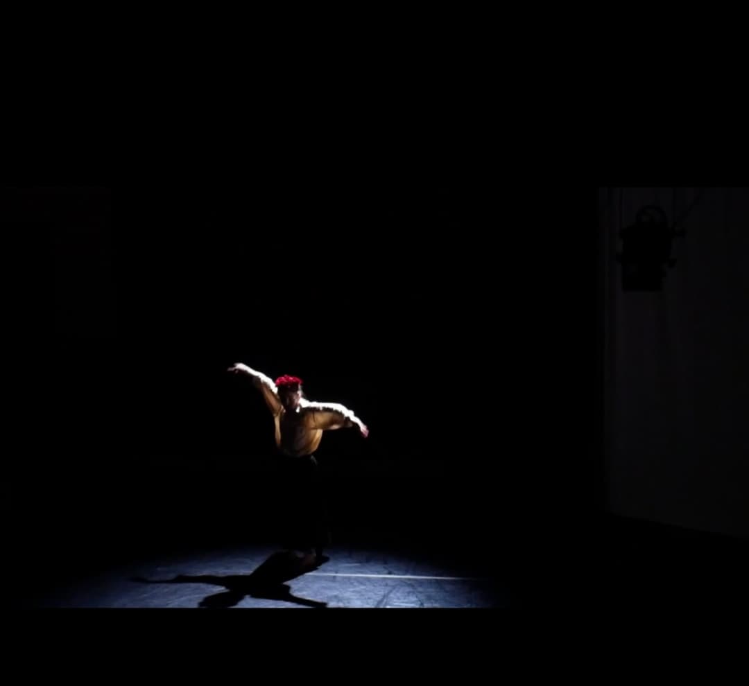 This image is very dark, and was captured of a dancer (myself) performing on stage lit by a warm, dim, soft spotlight. My arms are raised to the side, my right arm slightly higher than the left, and it was captured following a turn in a suspended position. I am wearing black, loose trousers, and a beige silky shirt tucked in.