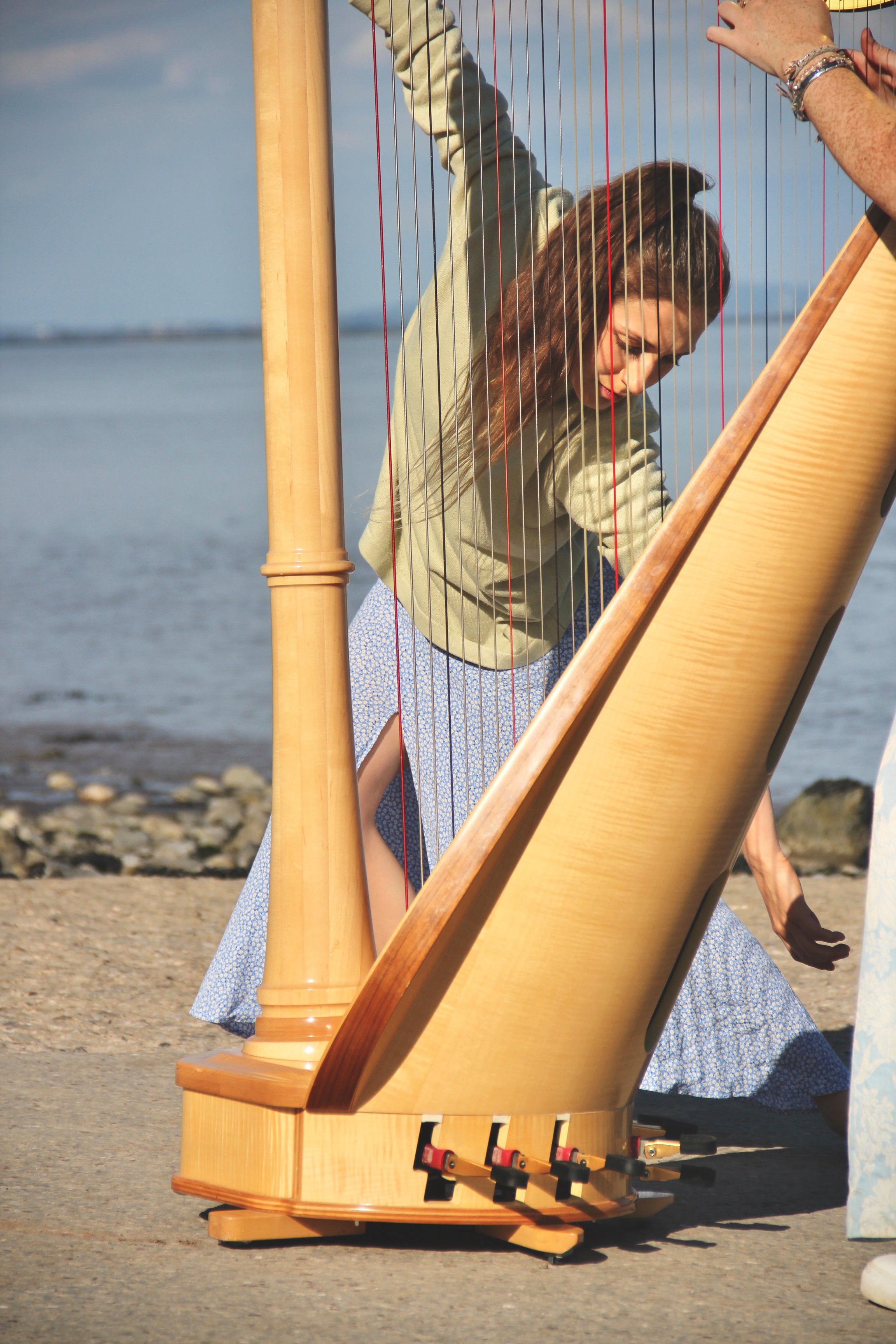 This image has been taken outside on a warm, sunny day by the sea. Most of the image is filled by a light wooden harp, and behind it is a dancer (myself) being captured in movement that can be seen through the frame of the harp. I am wearing a pastel green jumper, and a light blue flowing long length skirt. In the background you can see the sea. It is calm and vast.