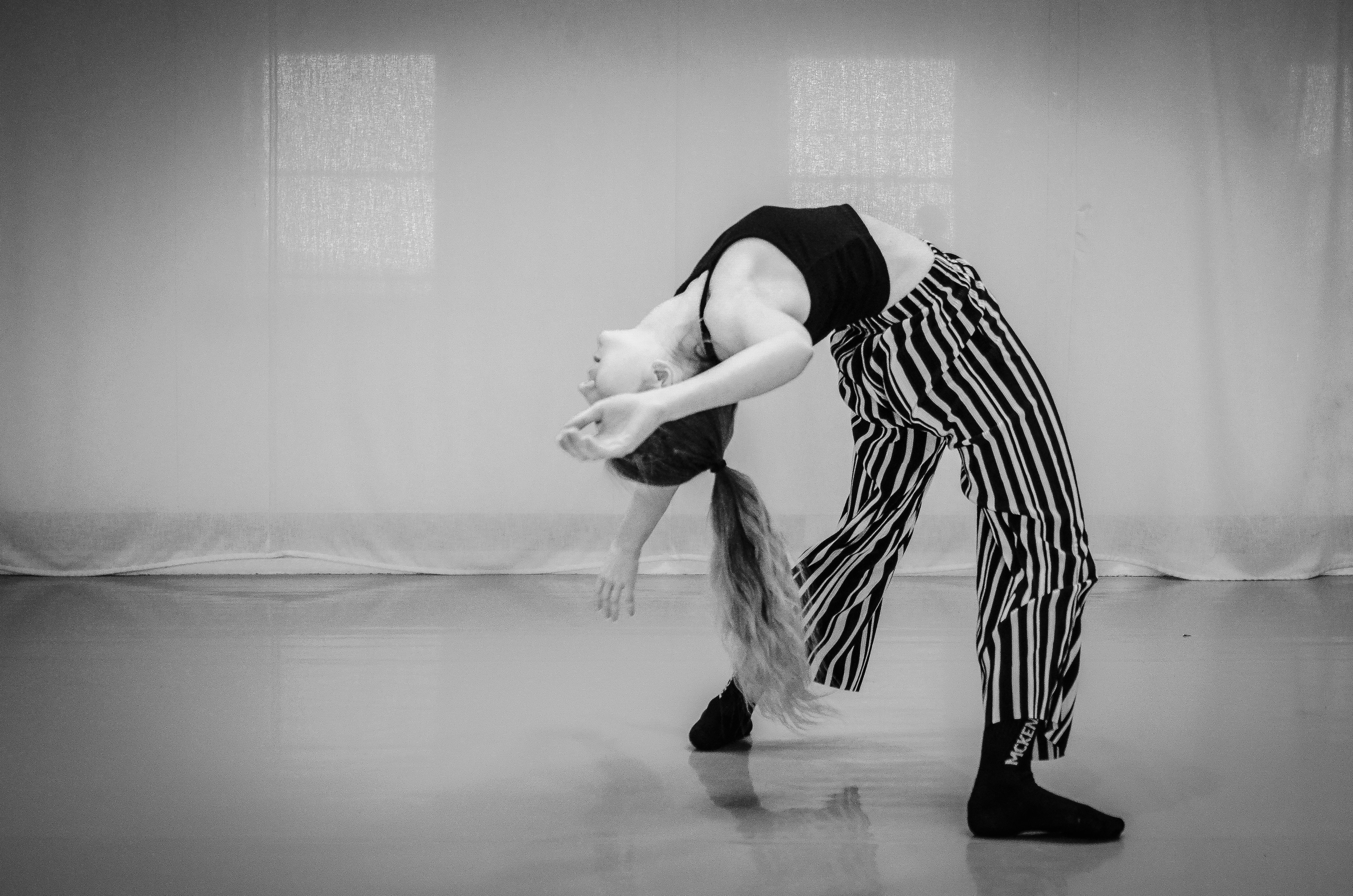 An image filtered in black and white, taken in a studio environment. The photograph captures a dancer (myself) in an arched position, with the arch in my back captured from the side. I am wearing striped trousers and a plain black vest top. My hair is long and held in a pony tail that is hanging towards the floor.