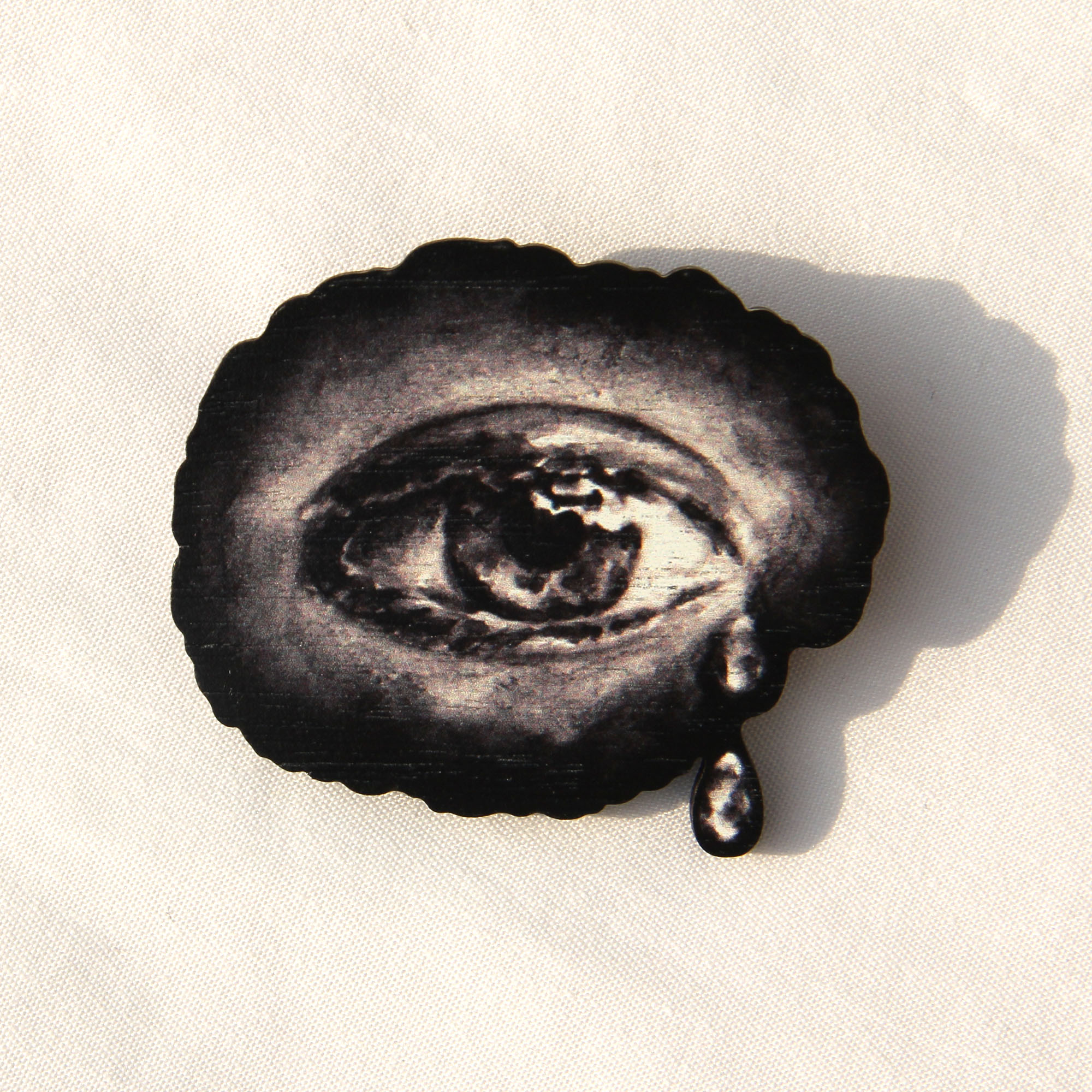 A wooden brooch printed with monochrome water-colour artwork of an eye with tears at the right corner on a white background.