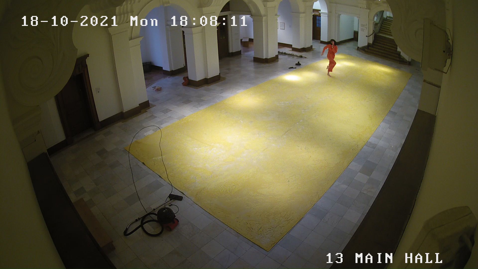 CCTV footage of performance at Glynn Vivian Gallery in Swansea. The image shows a figure dancing in an orange outfit on a yellow floor. 