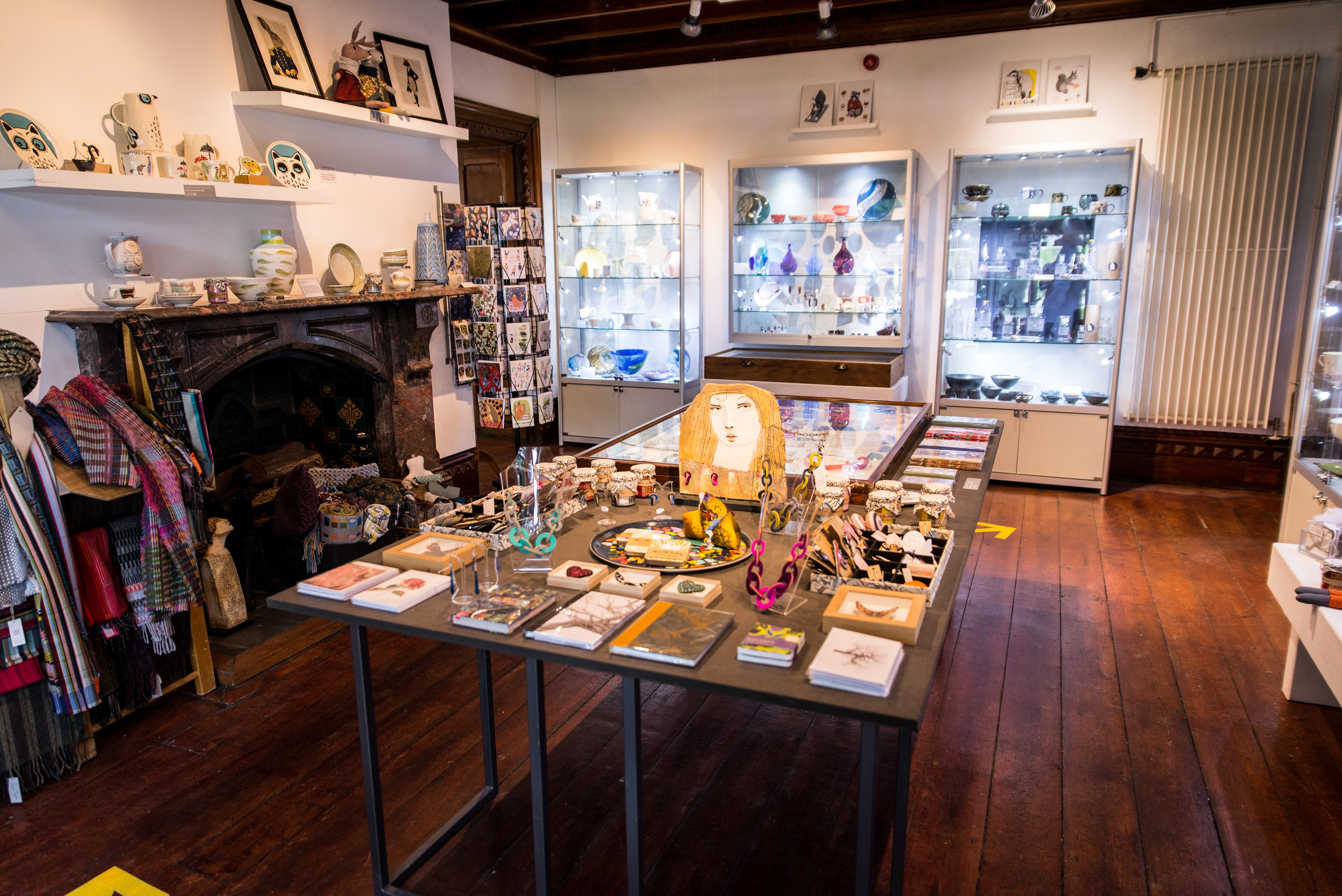 A view of our craft shop which has handmade items in glass cabinets and on tables. It also features an original fireplace and dark wooden floorboards.
