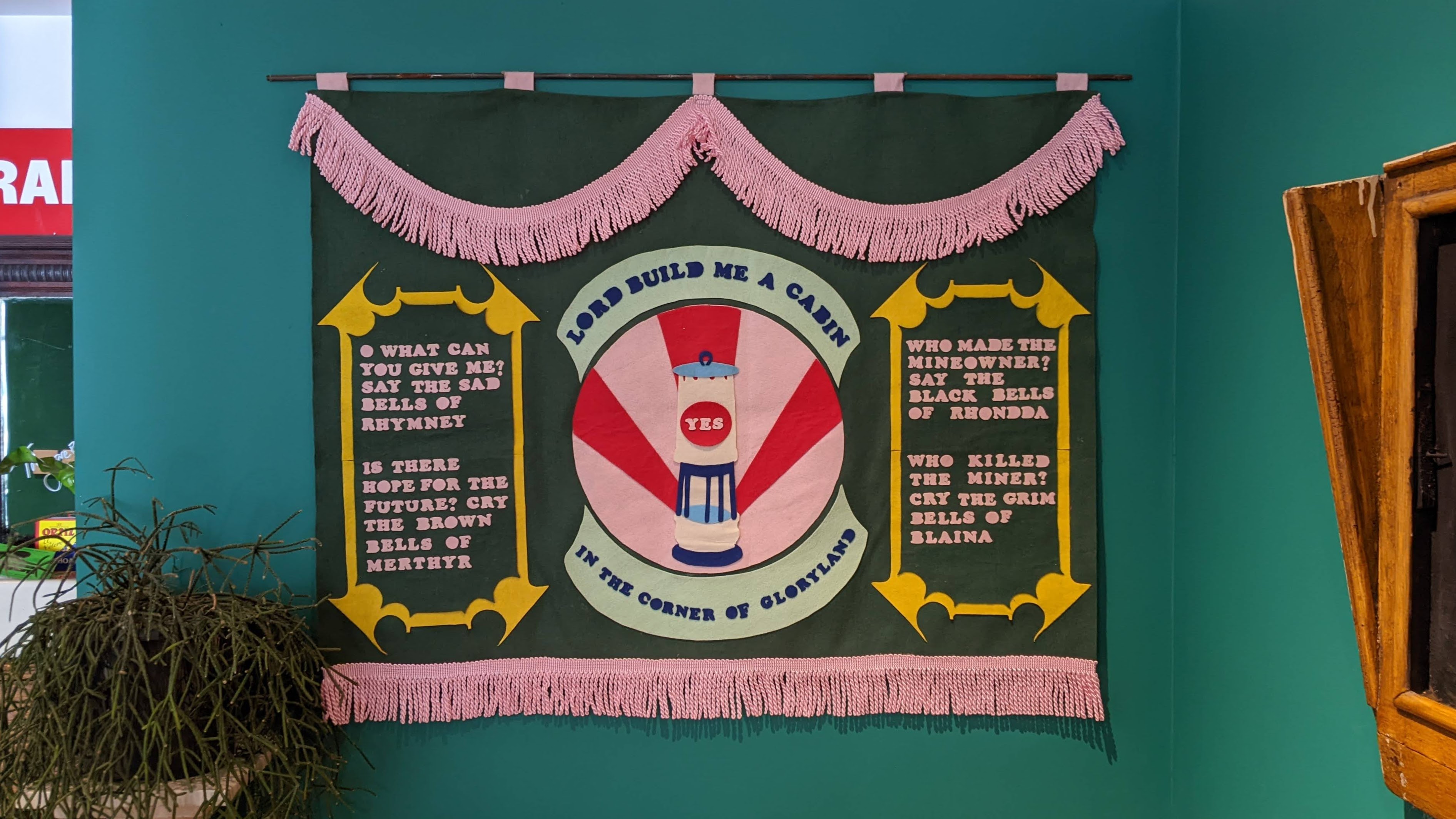 A forrest green banner with bright pink fringing and a colourful miners' lamp in the centre. Panels on each side display the words of Idris Davis' poem 'The Bells of Rhymney'