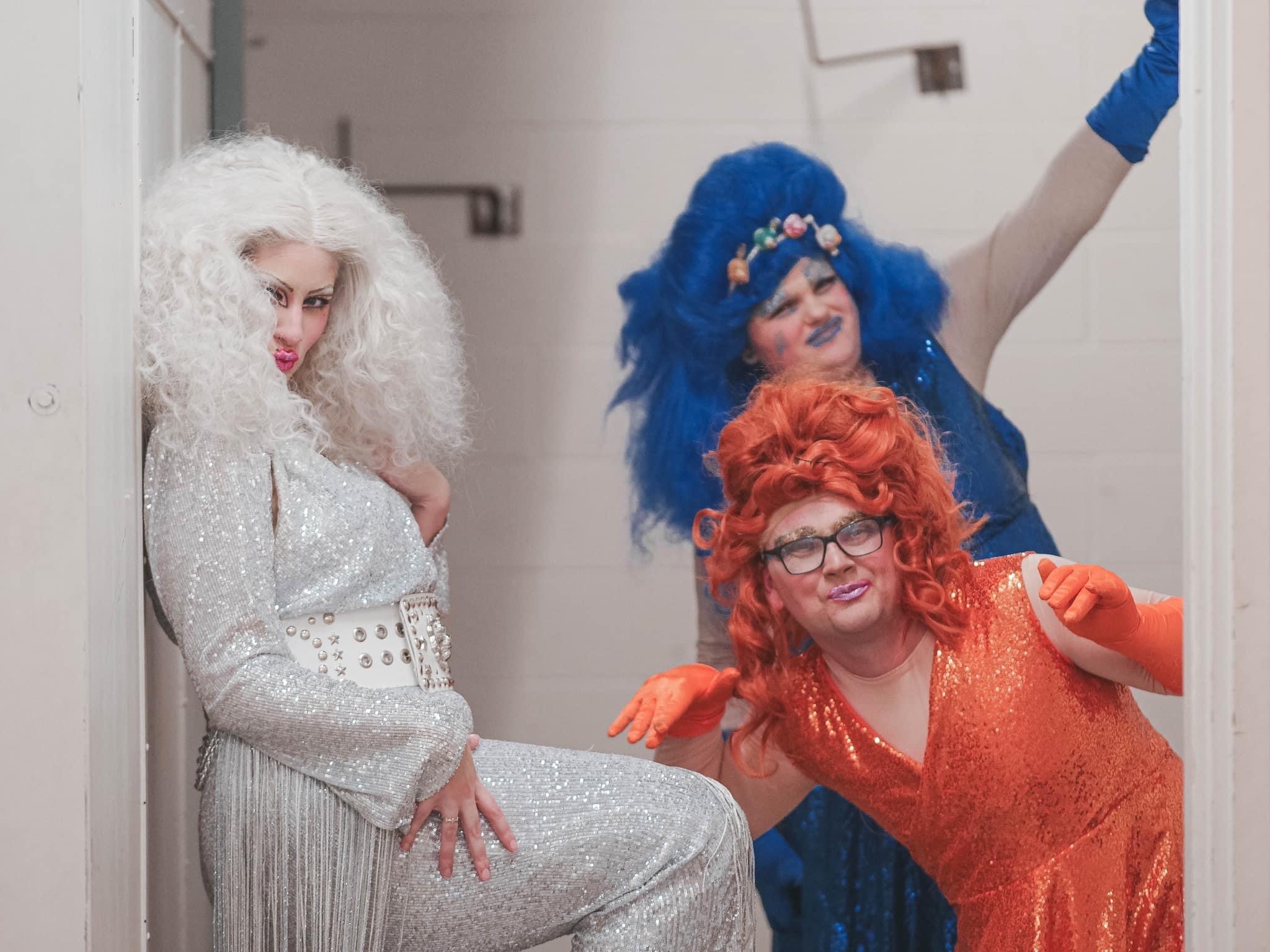 Three drag queens from the house of deviant. Cheesecake is wearing an orange sequin gown and wig, flossy is wearing a blue sequin gown and wig, and Cherry is in a silver sequin jumpsuit and wig. They are posing for the camera backstage.