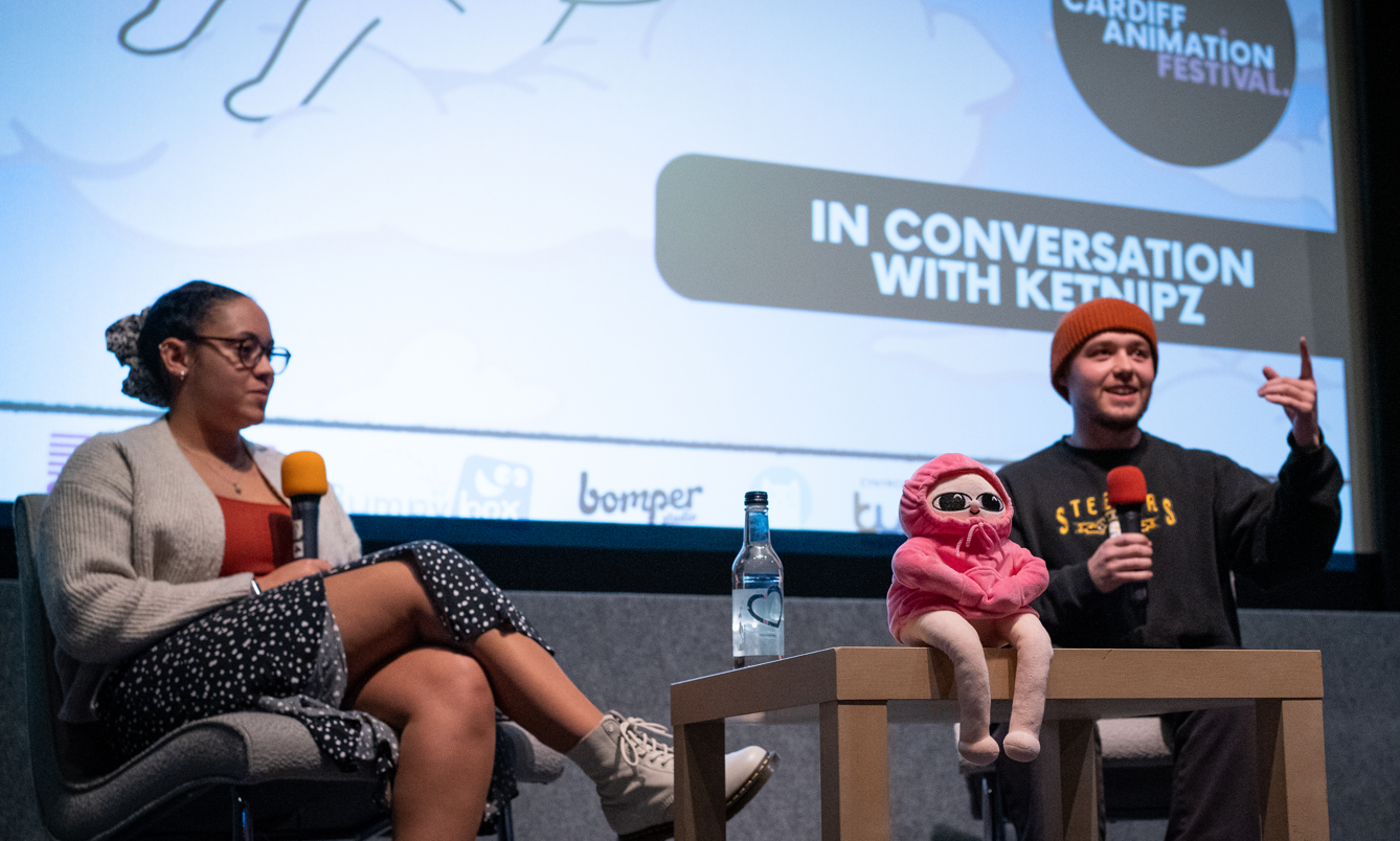 Erykah and Harry Hambley (Ketnipz) sat together sat together with a small coffee table in front of them, with the pink bean teddy sat on the table. Behind them is a holding slide that reads ' In Conversation with Ketnipz'