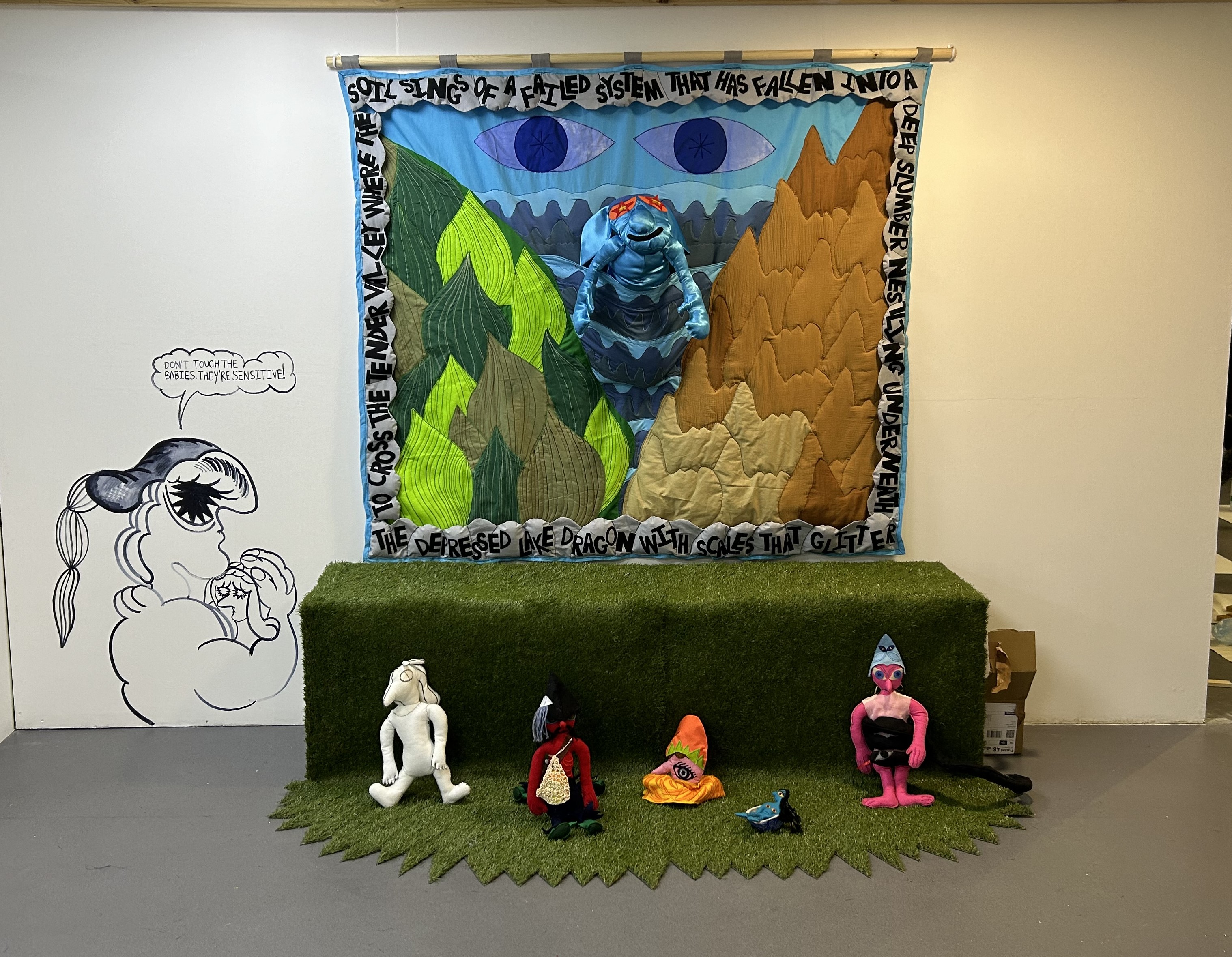 A mixed media piece, it includes: a tapestry that contains a lettered border that are taken from the artist's writing "To cross the Tender Valley where the soil sings of a failed system that has fallen into a deep slumber Nesting underneath the depressed lake dragon with scales that glitter and shaped like frozen sweat". The tapestry has a pocket in the middle which carries the depressed lake dragon, a character made from textiles. Below the tapestry, astroturf is placed underneath where four characters sit