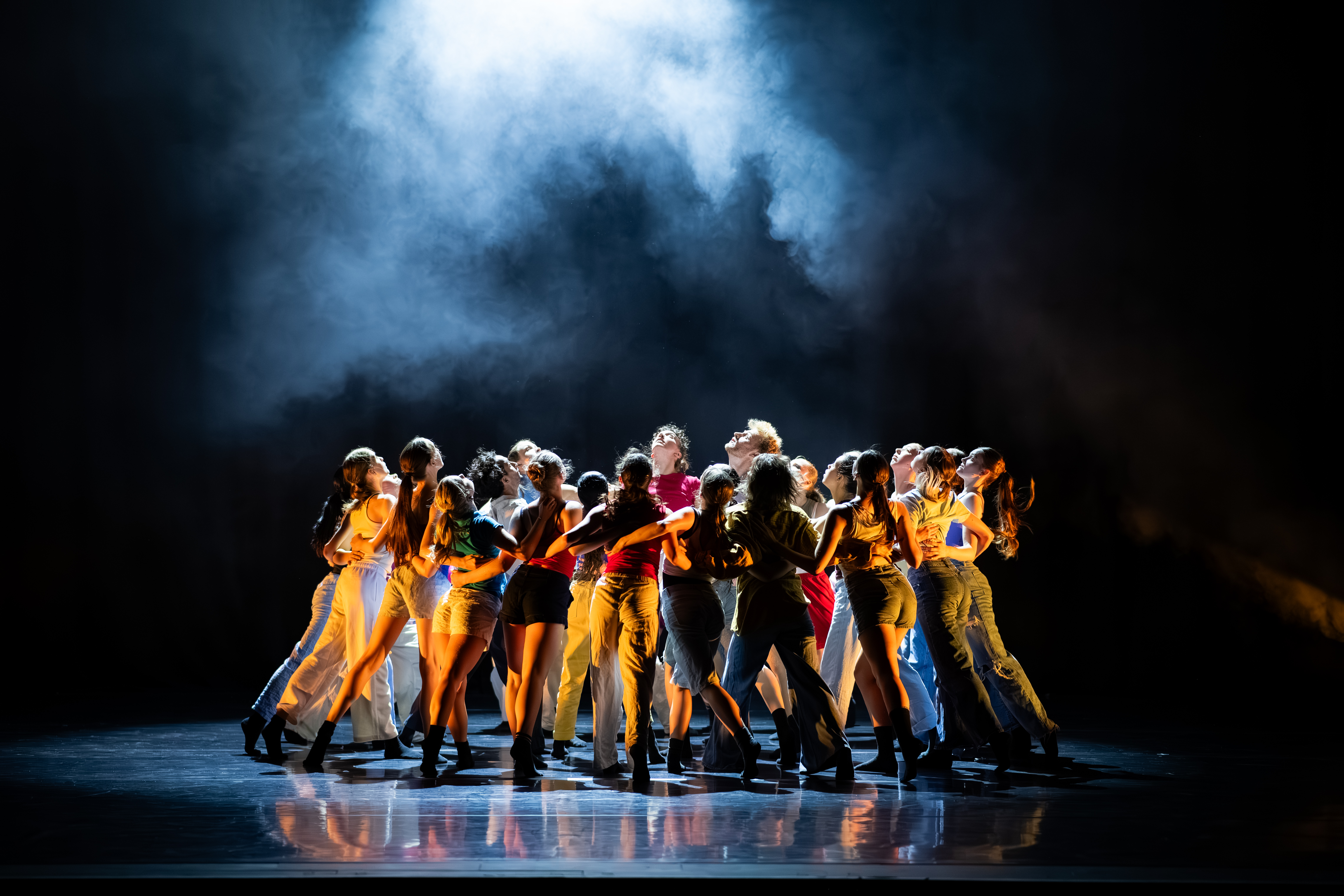 A cluster of Dancers, in a collective pedestrian clothing style, lit by warm golden side lights, looking up towards a cold cloud of haze