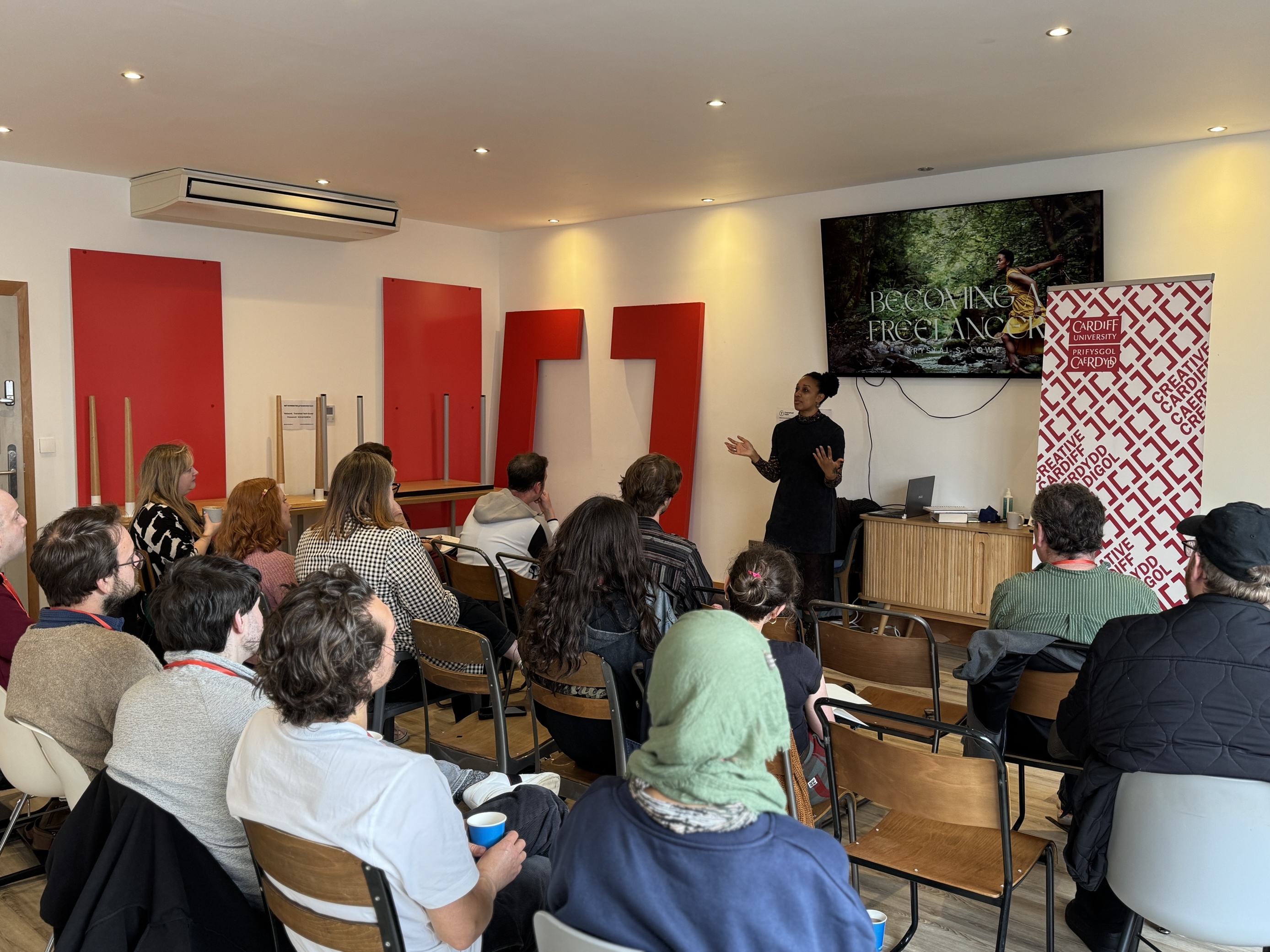 A speaker stands smiling at the front of a modern, bright room during a presentation titled ‘Becoming a freelancer.'
