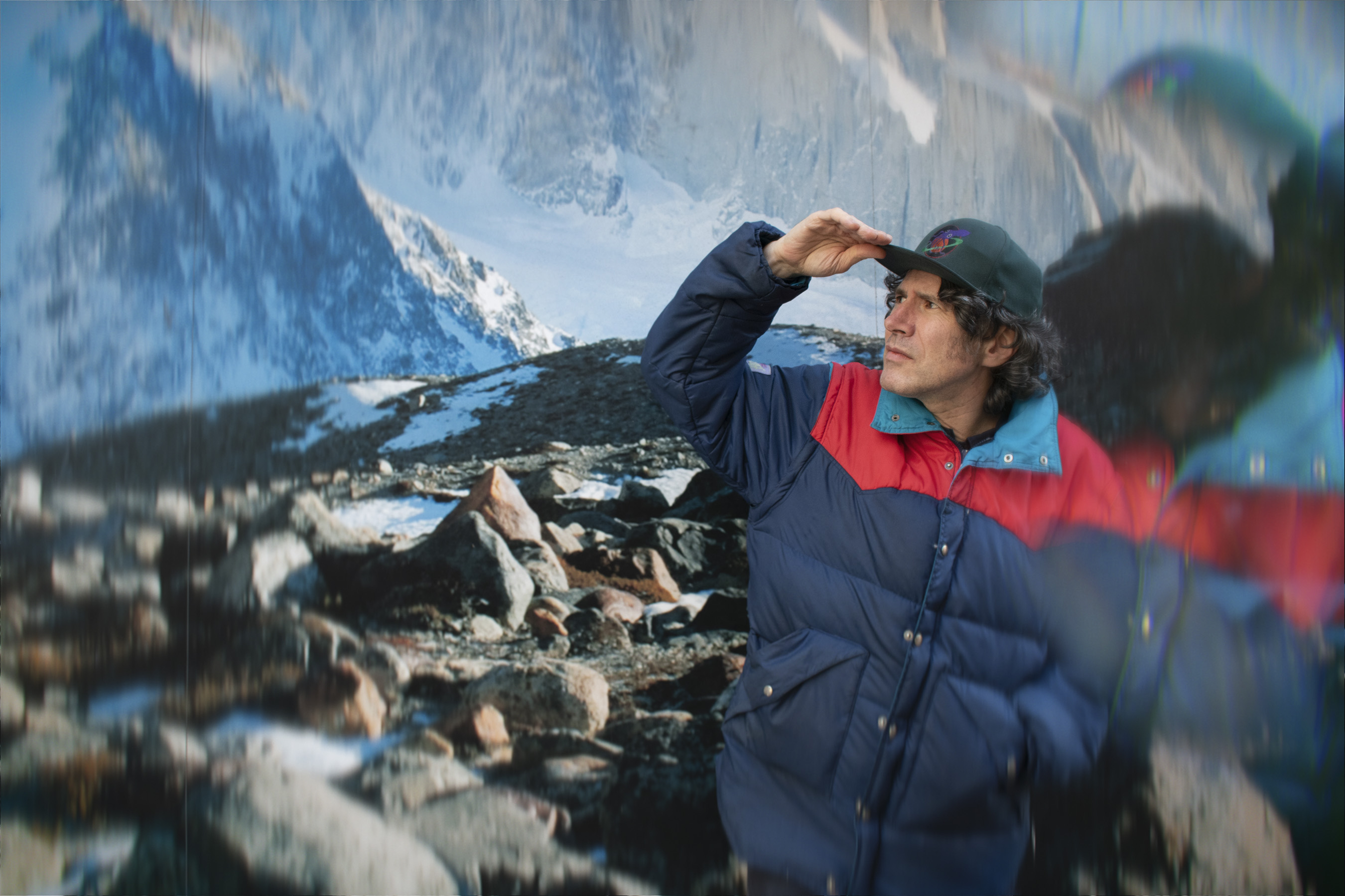 Gruff Rhys in blue jacket and cap against mountain backdrop