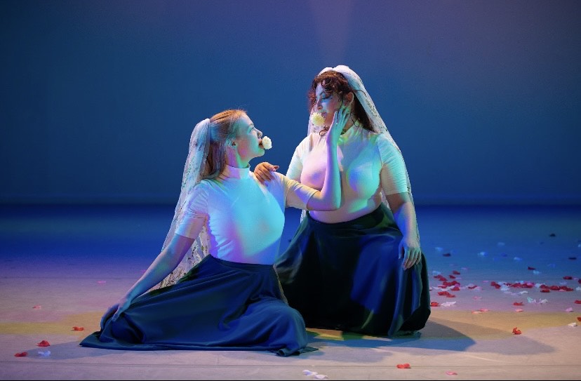 Two people on a blue lit stage next to eachother in costume, one is sitting the other crouching