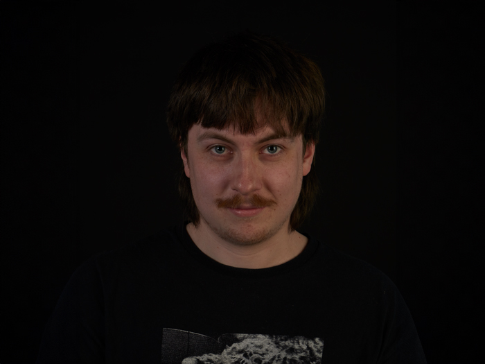 A person with a moustache stood in front of a black background