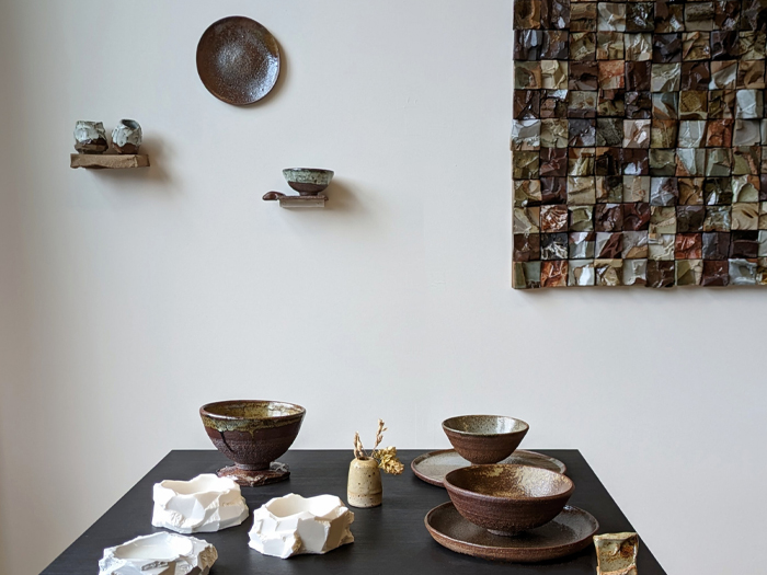 A white-cube gallery space with ceramic tiles, a plate and small vessels on the walls, and a table with bowls and plates on it