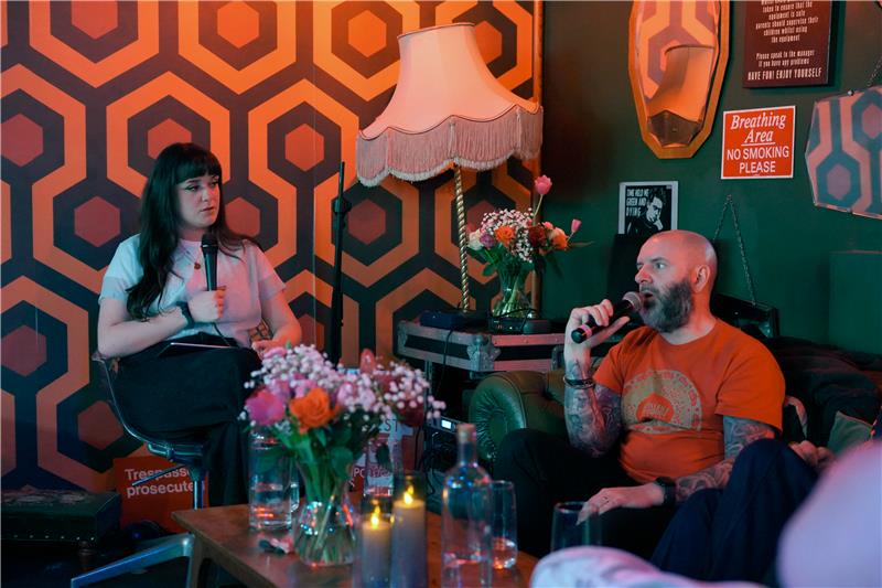 Tori sits holding a microphone opposite a man also holding a microphone mid conversation