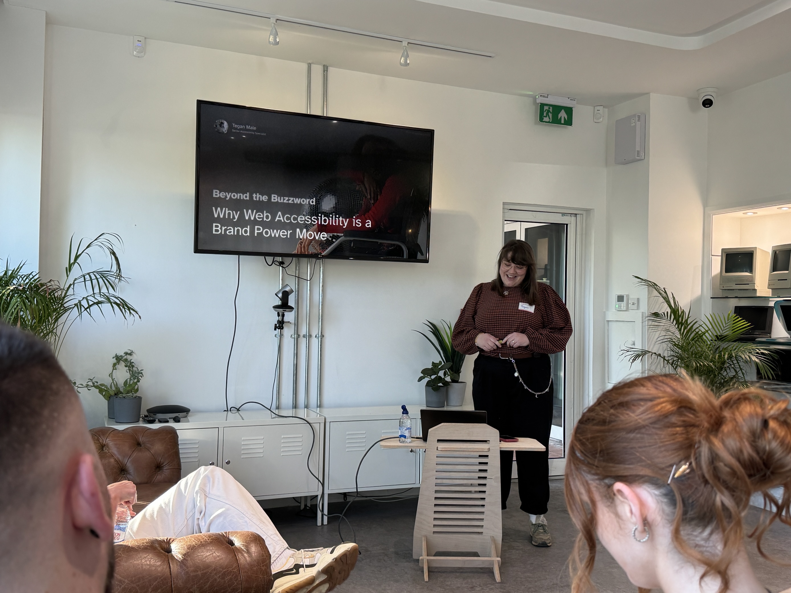A speaker stands smiling at the front of a modern, bright room during a presentation titled ‘Beyond the Buzzword: Why Web Accessibility is a Brand Power Move